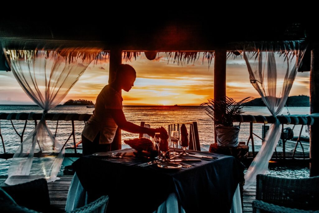 Staff member giving finishing touches to beautifully set open air restaurant table while the sun sets over the ocean views in the distance. 
